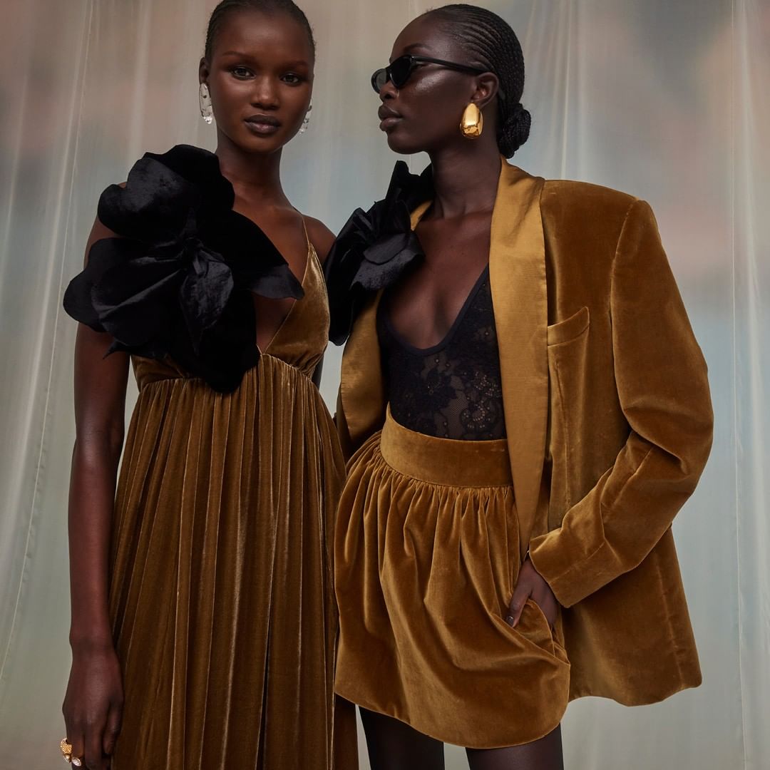 Two adult women pose in a studio wearing gold and black outfits with large floral accents. One is in a long dress, the other in a skirt suit.