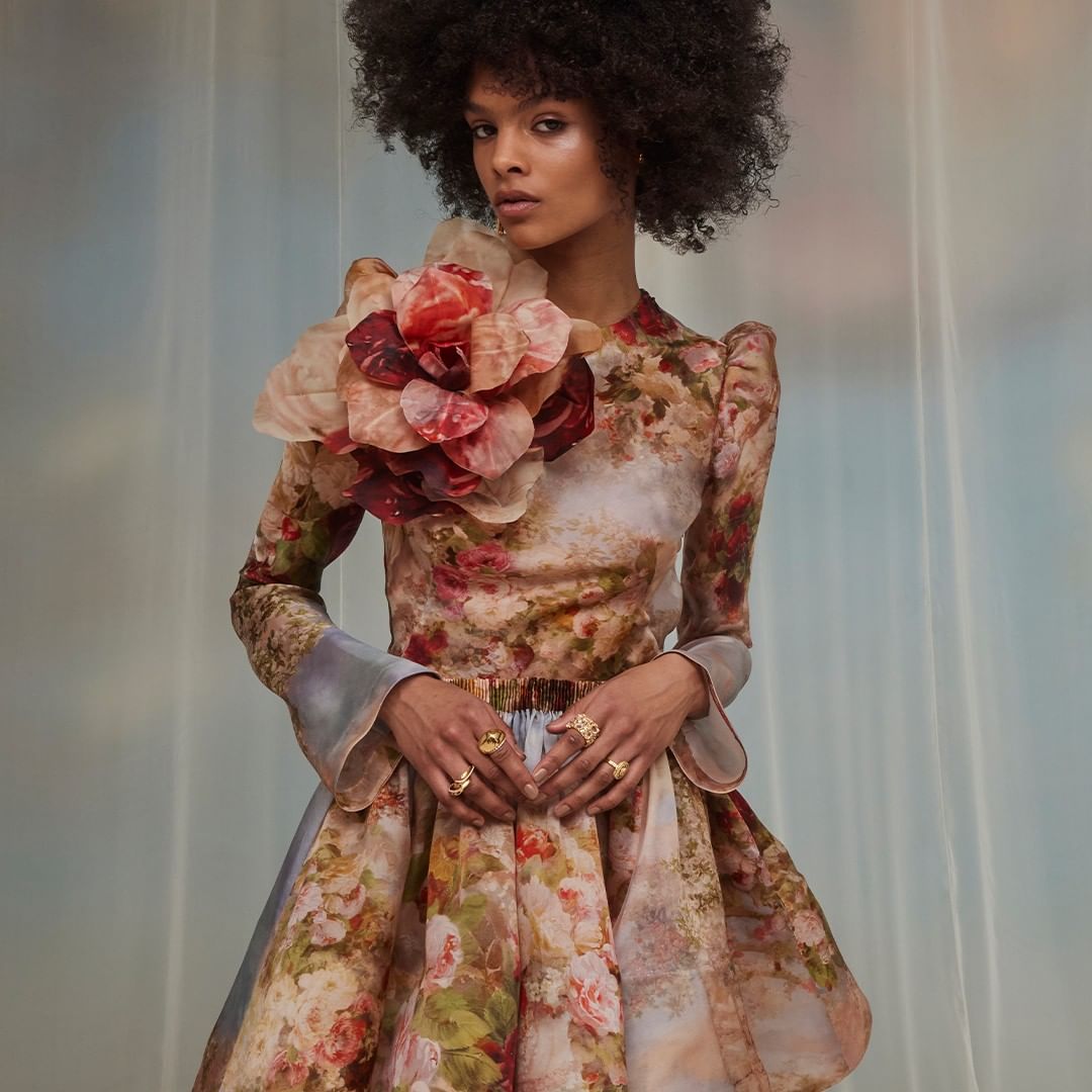 A woman in a floral dress stands indoors in a medium shot, showcasing a large flower on her shoulder and an elegant, stylish look.