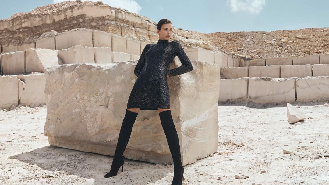 Fashion shot of woman posing against large marble blocks. She wears a dark dress and thigh-high boots in bright outdoor light.