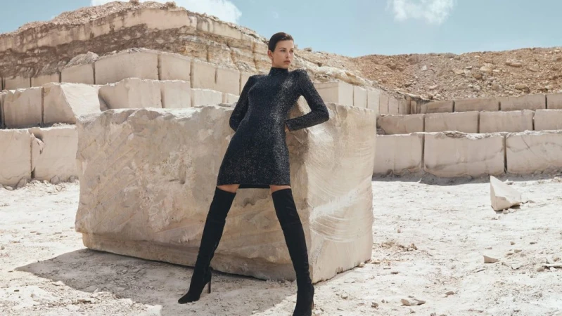 Fashion shot of woman posing against large marble blocks. She wears a dark dress and thigh-high boots in bright outdoor light.