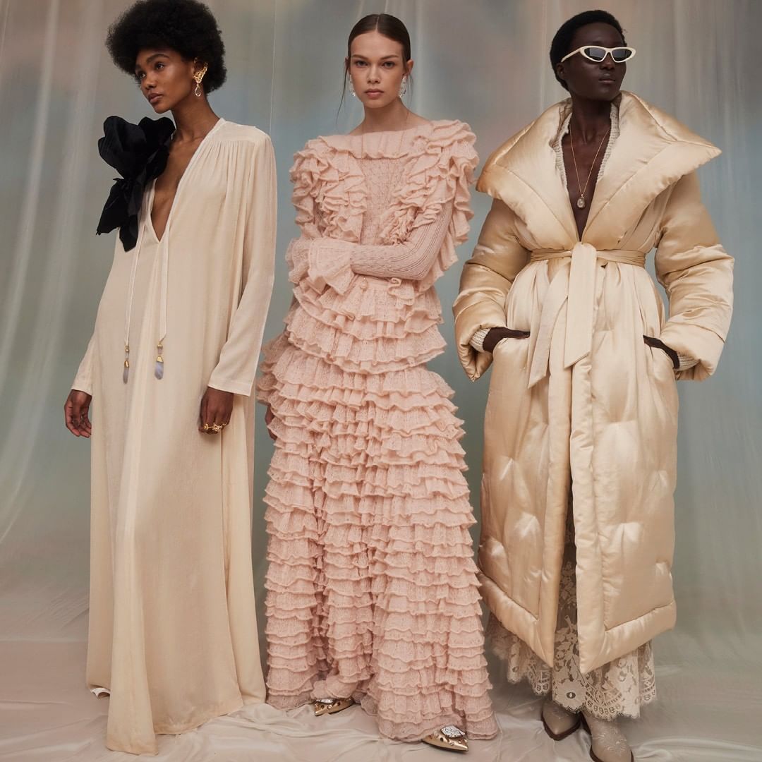 Three fashion models in long designer dresses strike a pose in a studio. One dress features ruffles, another a large black flower.