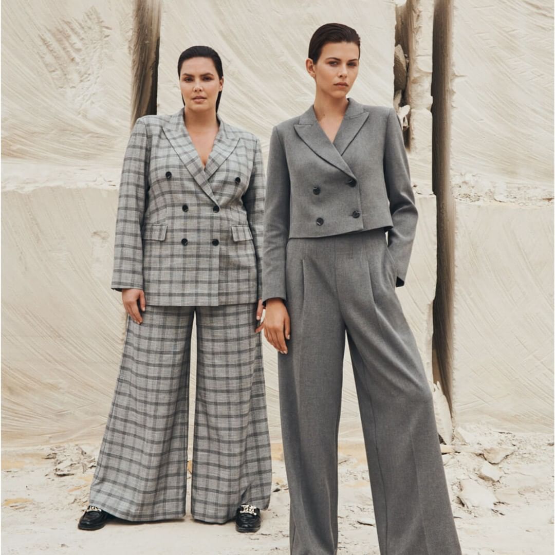 Two women wearing gray pantsuits pose in front of a textured stone background. One suit is plaid, the other is solid gray.