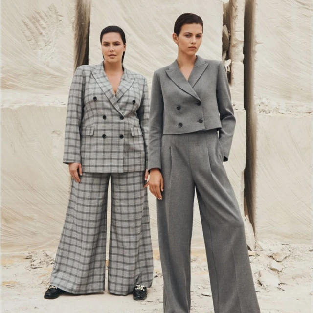 Two women wearing gray pantsuits pose in front of a textured stone background. One suit is plaid, the other is solid gray.
