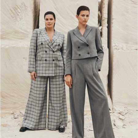 Two women wearing gray pantsuits pose in front of a textured stone background. One suit is plaid, the other is solid gray.