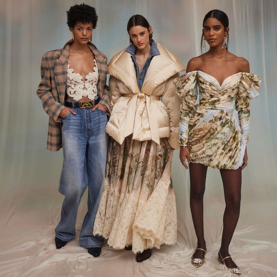 Three fashion models pose indoors in a studio setting. One wears a blazer, jeans, and a white top. The others wear dresses and outerwear.