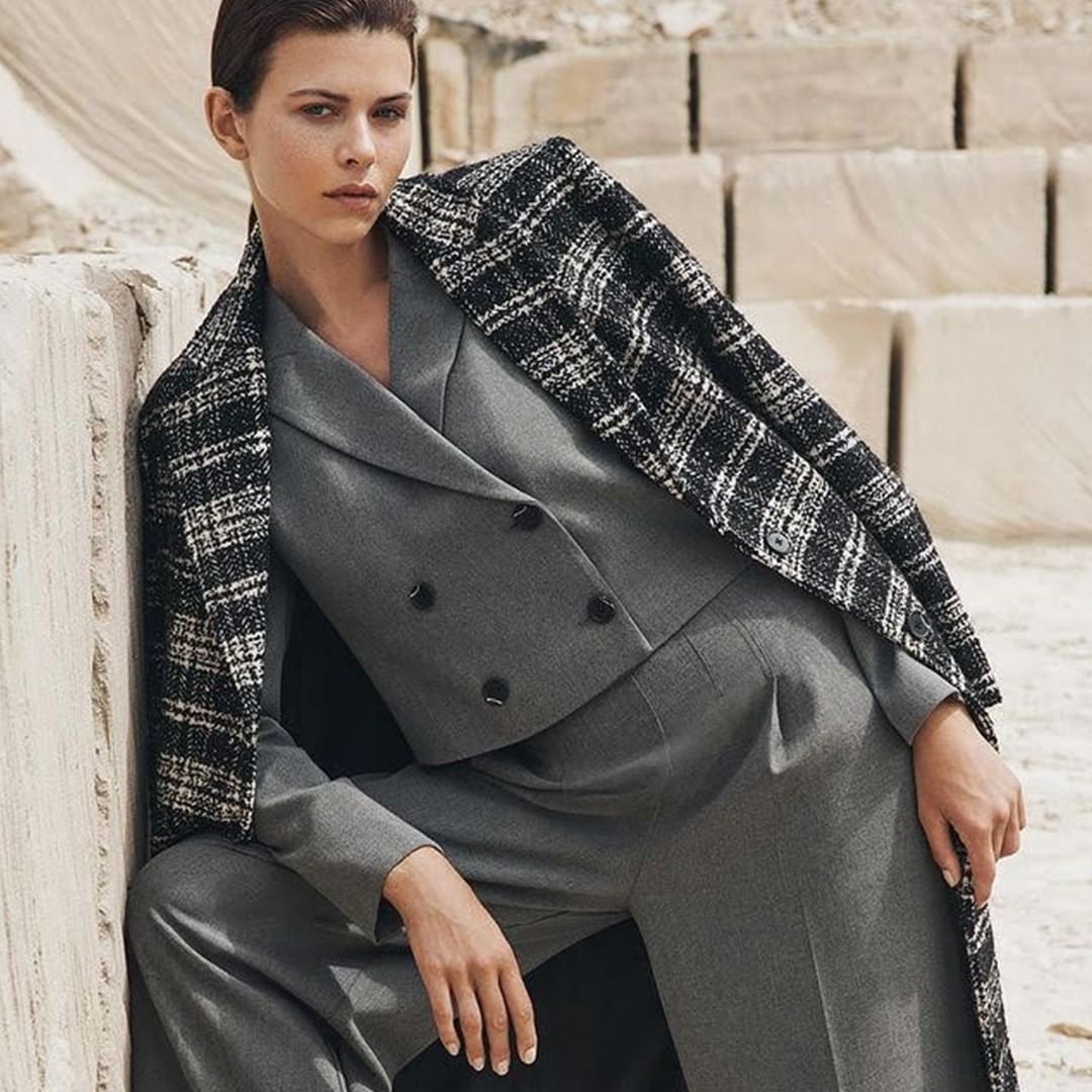 A young woman wearing a gray suit and black and white plaid coat poses in front of stacked stone blocks outdoors, fashion shot.