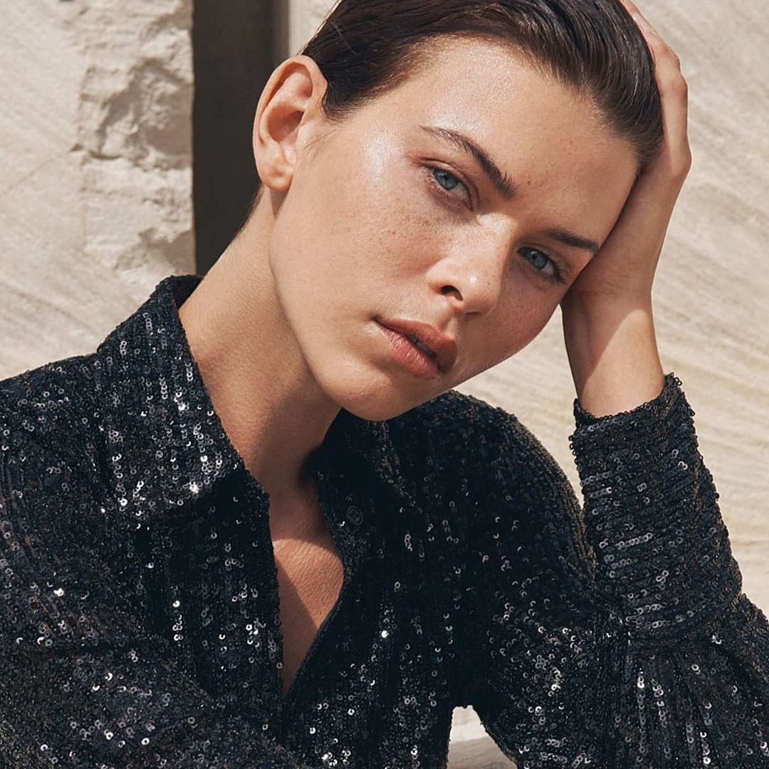 Close-up portrait of an adult female model with fair skin and blue eyes wearing a sparkling black button-down shirt against a stone backdrop.