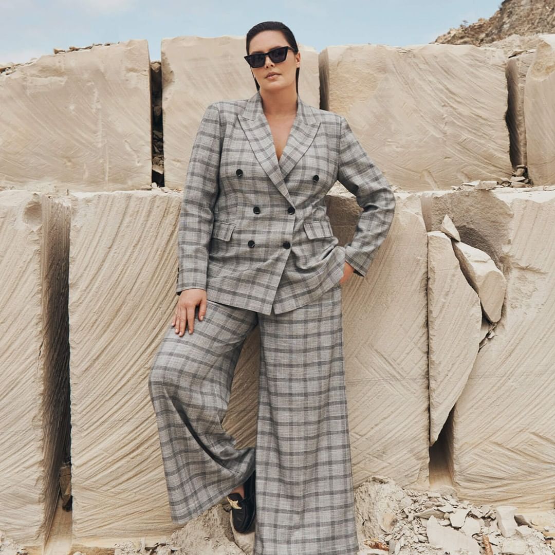 Adult female fashion model in a gray plaid suit poses outdoors against a background of large, pale stone blocks in daylight.