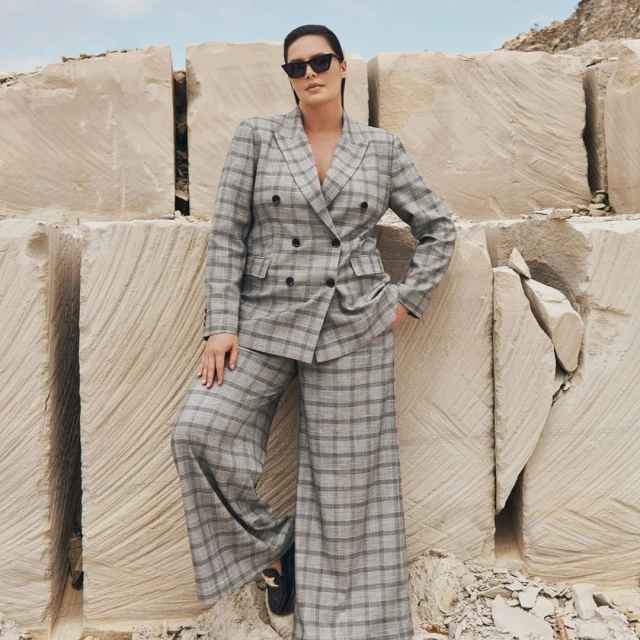 Adult female fashion model in a gray plaid suit poses outdoors against a background of large, pale stone blocks in daylight.