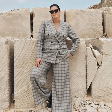 Model wearing a grey plaid pantsuit and dark sunglasses standing against large stacked sandstone blocks in Sydney.