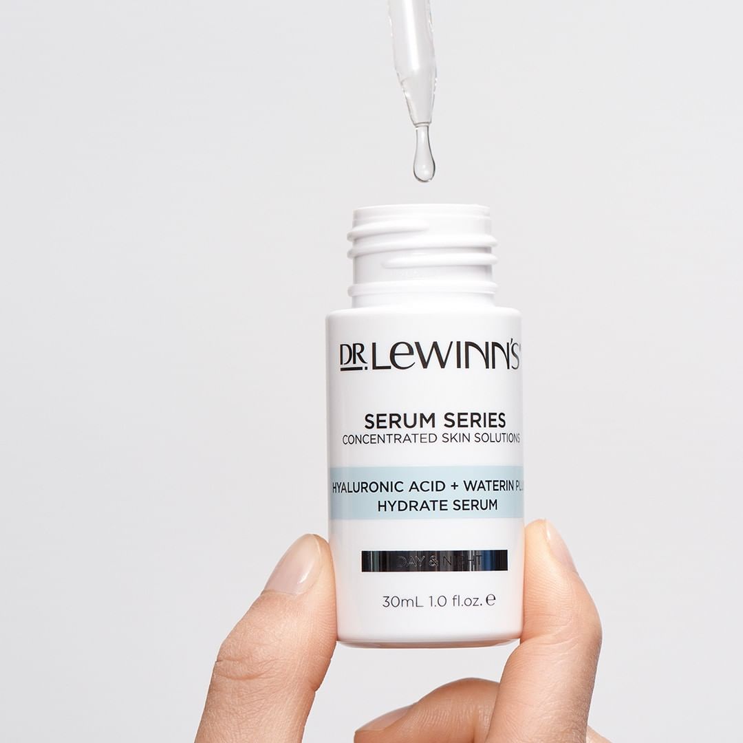 Close-up of a hand holding a white bottle of Dr. Lewinn's serum with a dropper dispensing liquid against a white background.