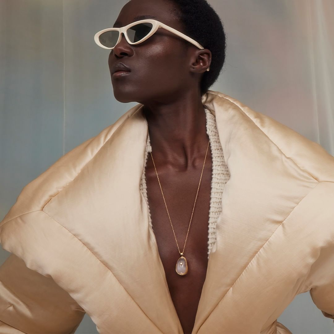 Fashion shot of a Black woman wearing white cat-eye sunglasses and a beige coat with a stone pendant necklace in front of a muted backdrop.