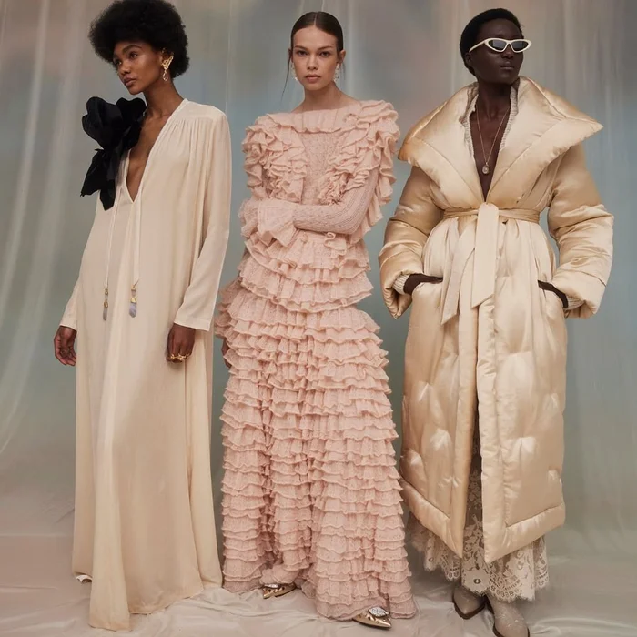 Three female models posing in cream and pink designer dresses and puffer coat against a fabric backdrop.