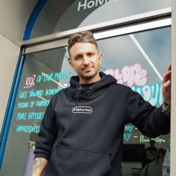 Man wearing a black hoodie standing in a doorway with blue and pink handwritten text on the glass behind him in Sydney.