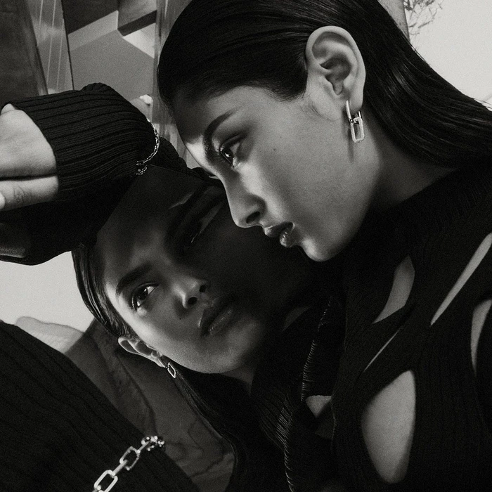 Monochrome close-up portrait of a woman wearing a black cutout top and linked silver earrings reflecting in a mirror surface.