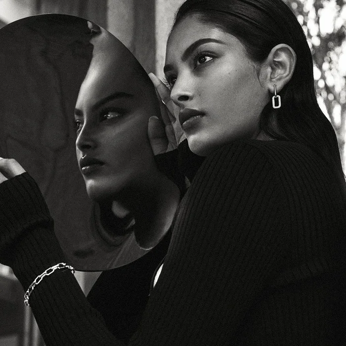 Monochrome portrait of a woman holding a curved round mirror reflecting her face, wearing a ribbed black top and silver jewelry.