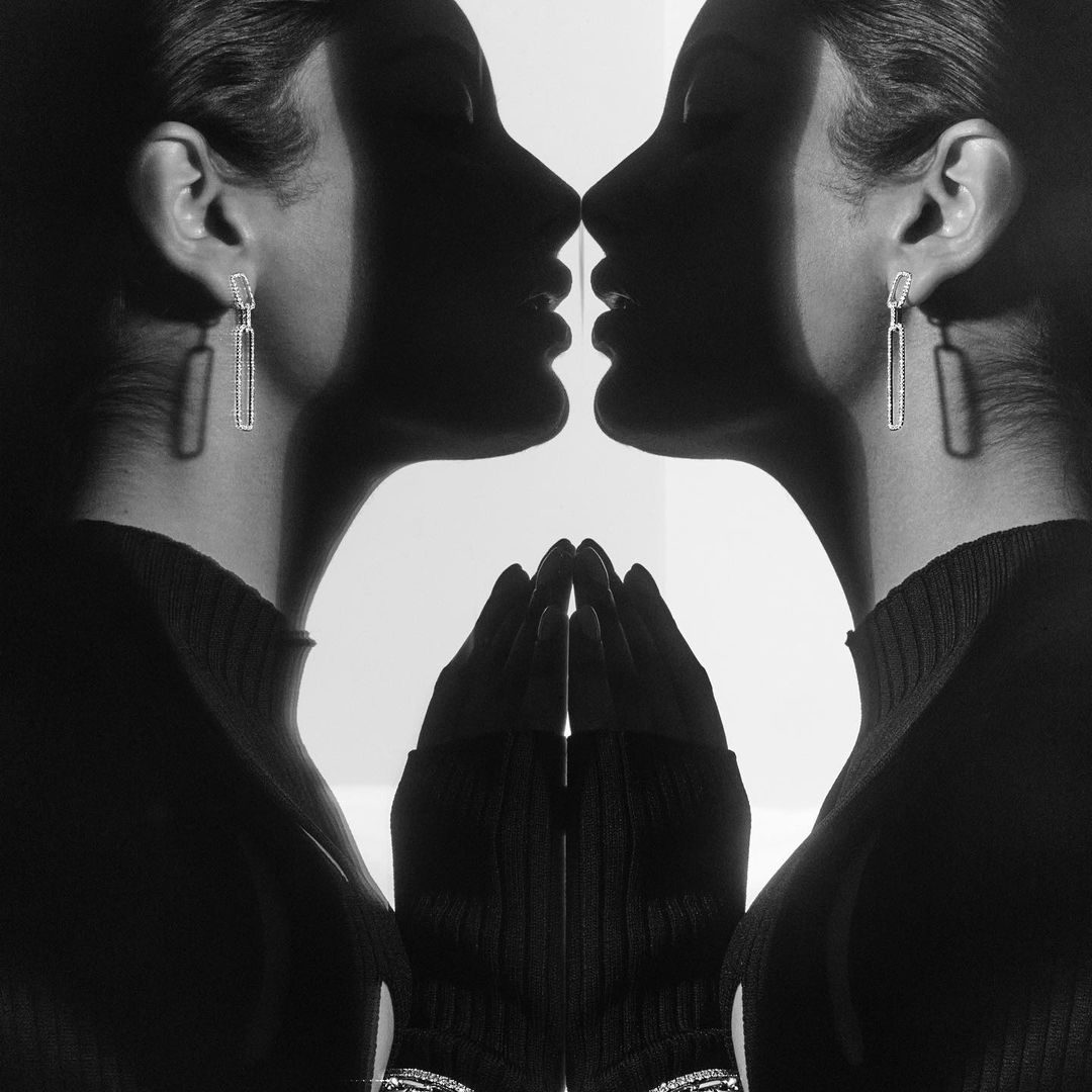 Monochrome portrait of a woman in a high-neck sweater and large earrings, mirrored, with hands near the face, against a white background.
