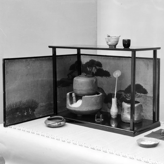 Japanese tea ceremony setup with ceramic bowls, a vase with sticks, and a painted screen on a display shelf, creating an elegant still life.