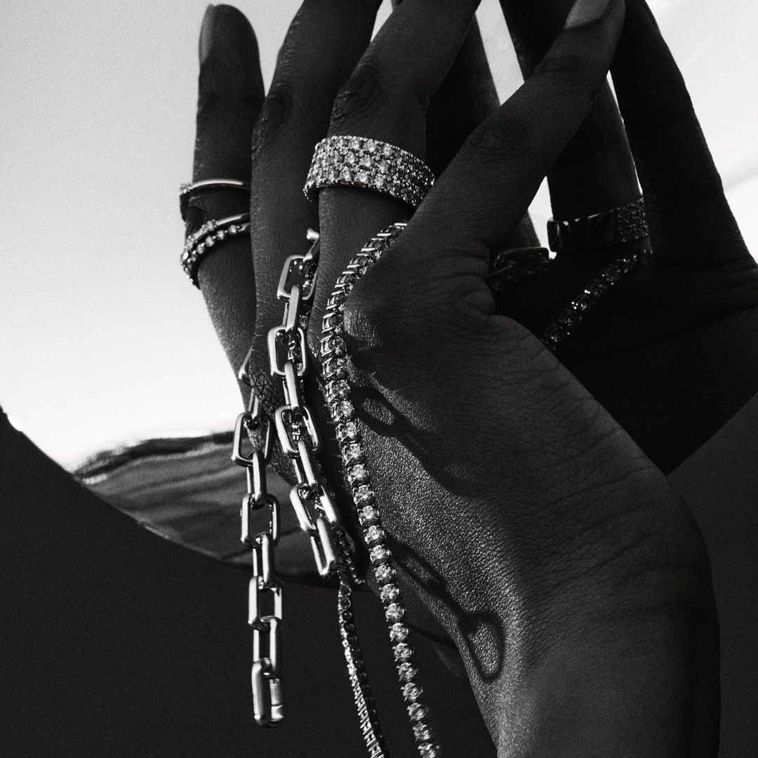 Close-up of a dark-skinned hand adorned with multiple rings and draped with silver necklaces against a bright, neutral background.