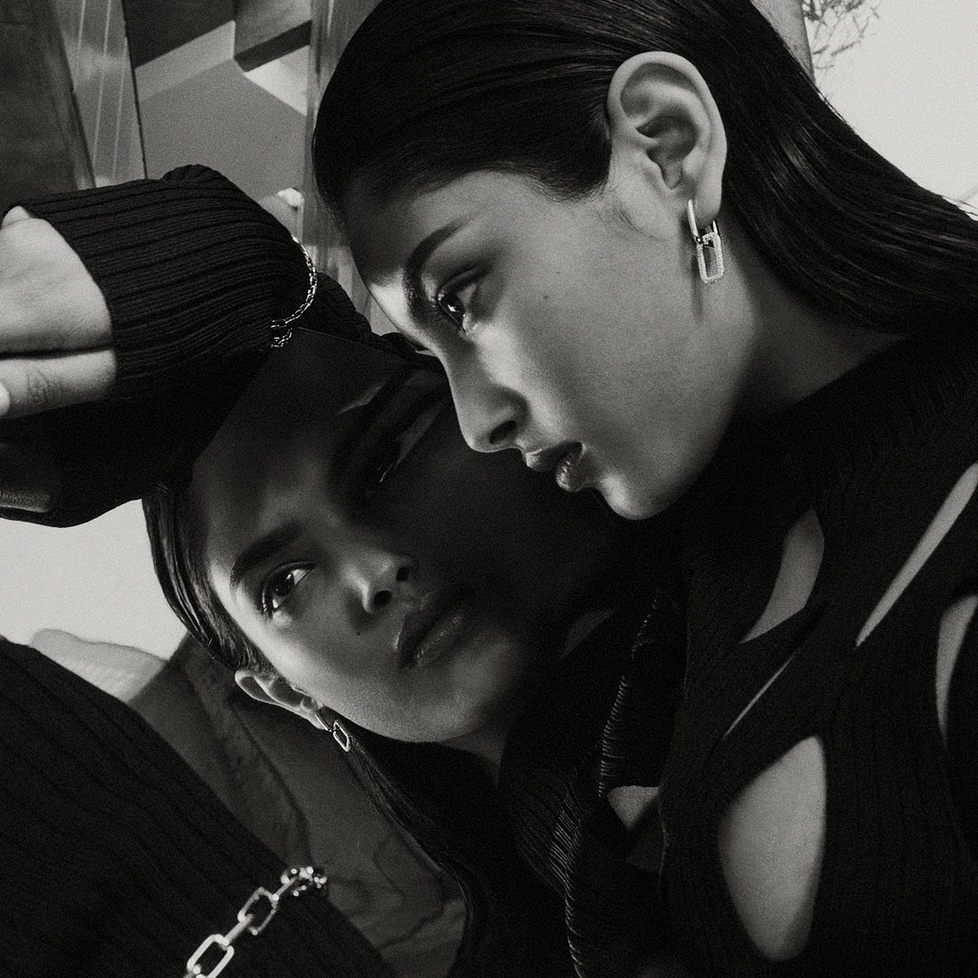 Two adult women pose in a close-up, monochrome portrait with sleek dark hair, contemporary earrings and clothing with cutout details.