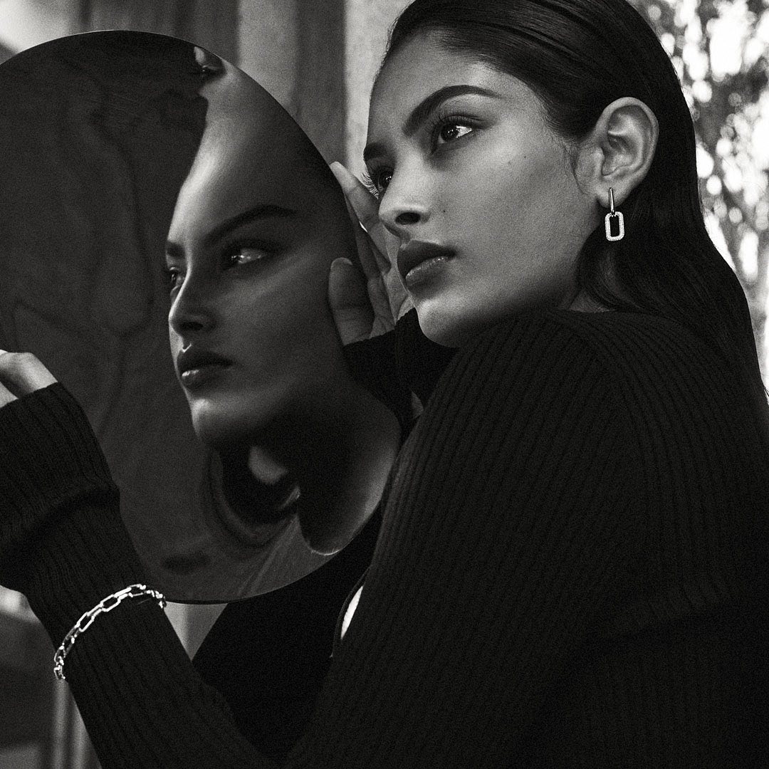 Monochrome studio shot of a woman in a black sweater wearing jewelry looking at her reflection in a mirror, and nature background.