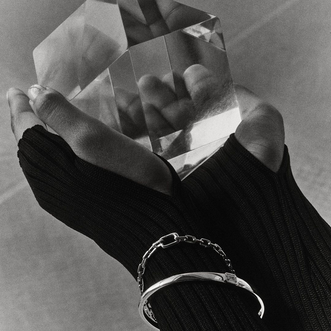 Elegant studio shot features a fair-skinned hand in black knit glove holding a clear geometric object, adorned with silver bracelets.
