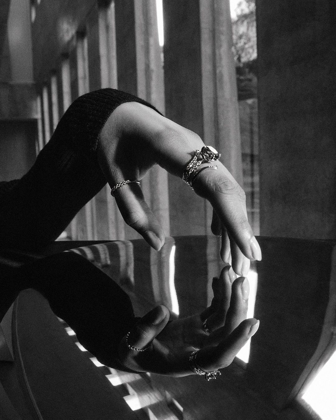 Close-up of two hands adorned with rings, reflecting each other, set against a backdrop of vertical architectural elements in black and white.