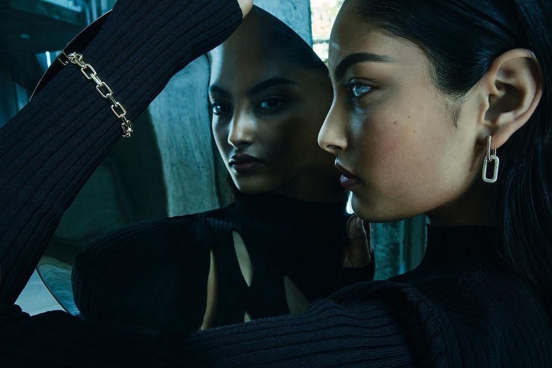 Woman's face and reflection, wearing a black top and silver bracelet and earrings, against a blue background, studio fashion shot.