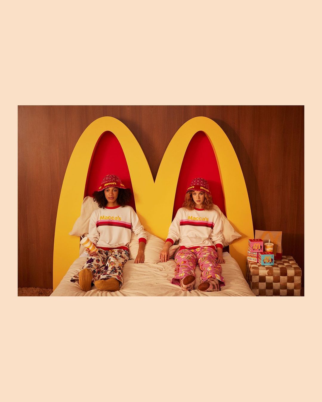 Two women sit on a bed in matching outfits in front of a McDonald's-themed headboard with additional branded objects.