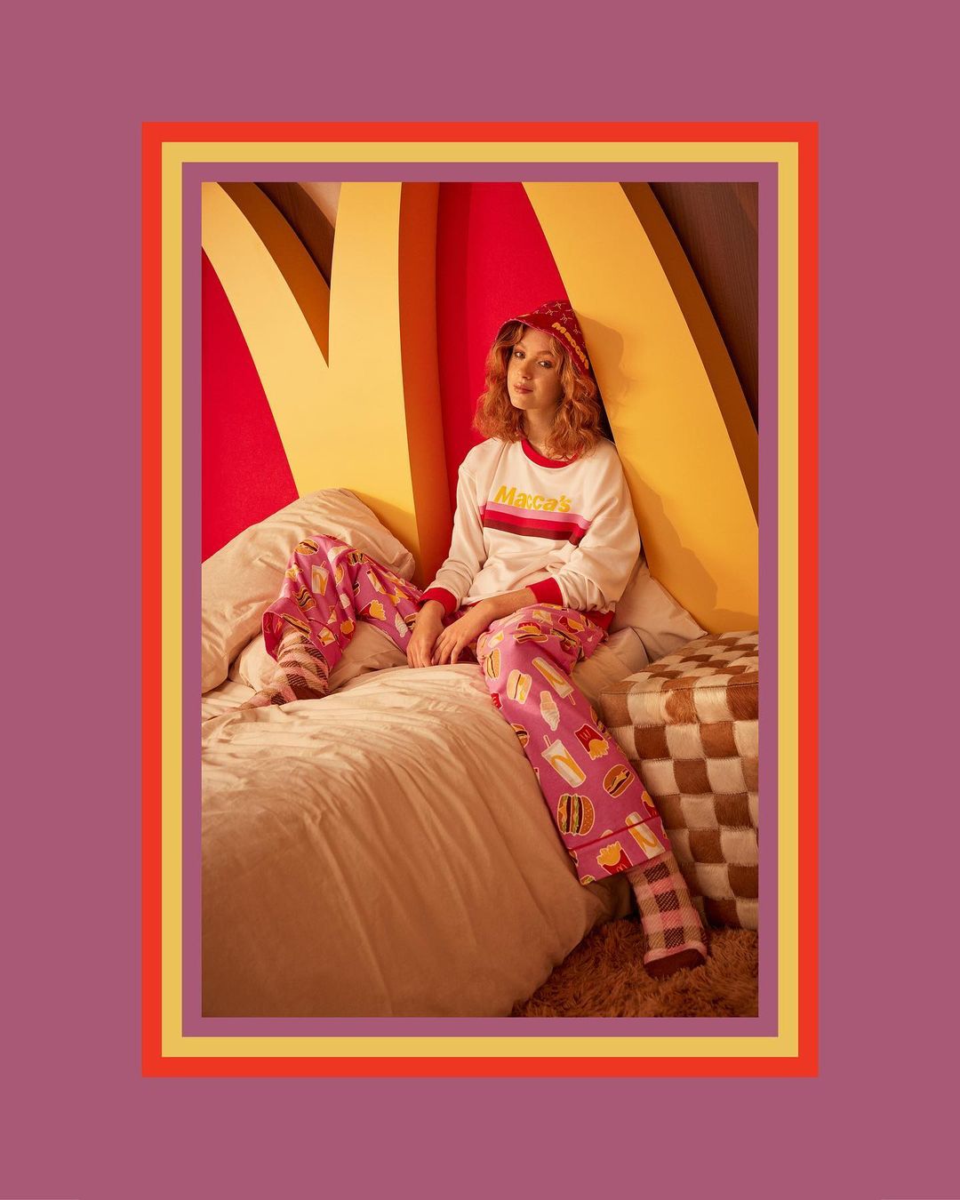 A redhead woman in pajama pants sits on a bed in front of yellow arch decor, creating a playful and inviting scene.