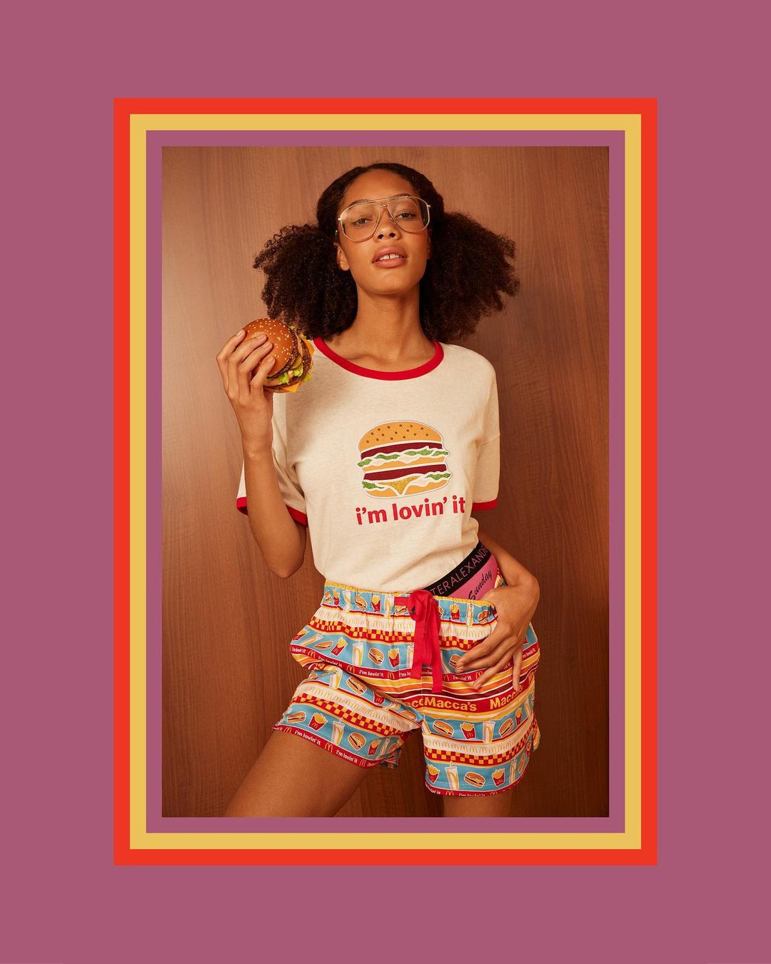 Young adult female wearing patterned sleep shorts and a graphic tee poses in an indoor setting while holding a burger.