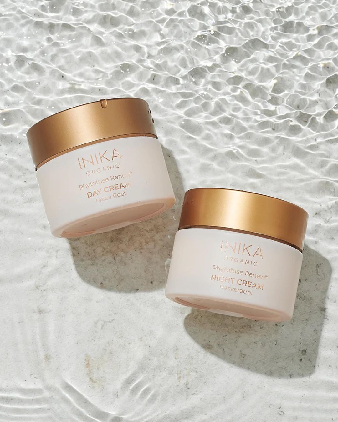 Two Inika Organic cream jars, a day and night cream, sit on a light marble surface with water ripples, emphasizing beauty product hydration.