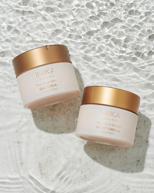 Two Inika Organic cream jars, a day and night cream, sit on a light marble surface with water ripples, emphasizing beauty product hydration.
