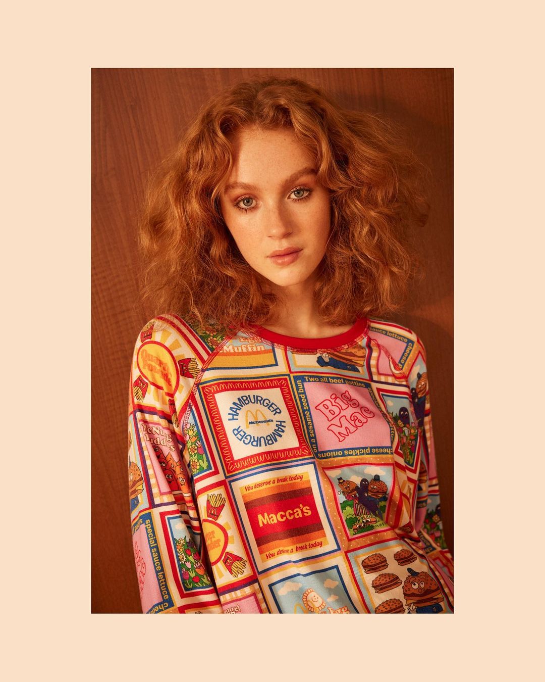 Portrait of a red-haired young woman wearing a colorful long-sleeve shirt with fast food graphics, set against a wood background.