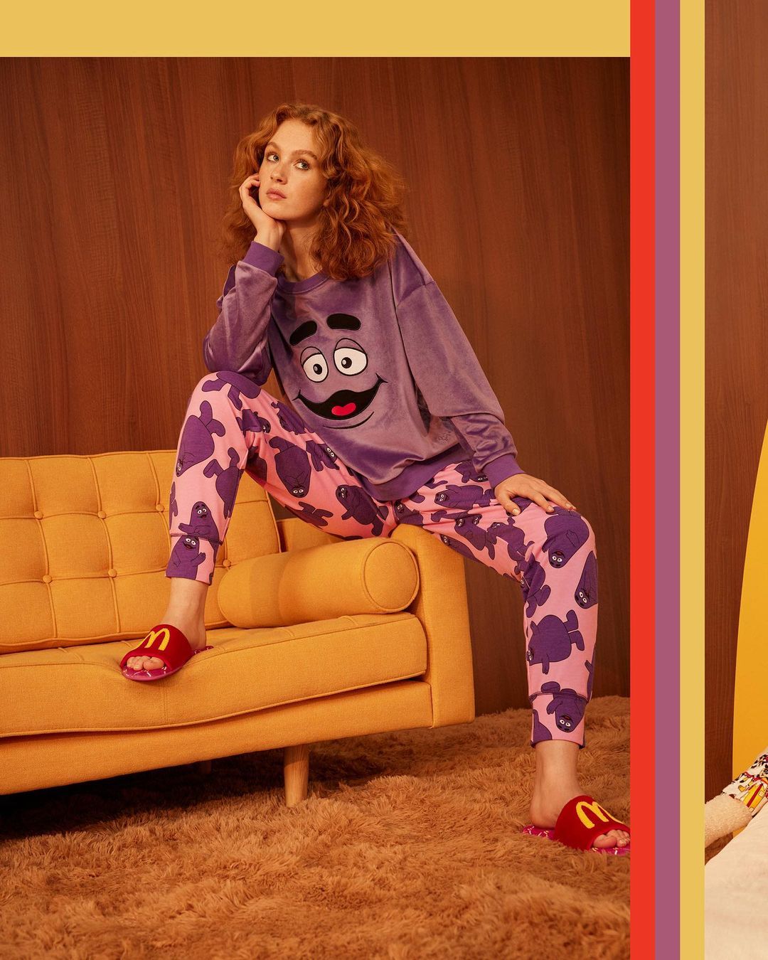 Woman in a purple sweatshirt with a cartoon face and patterned pants sitting on a yellow couch with red slippers in a warm-toned interior.