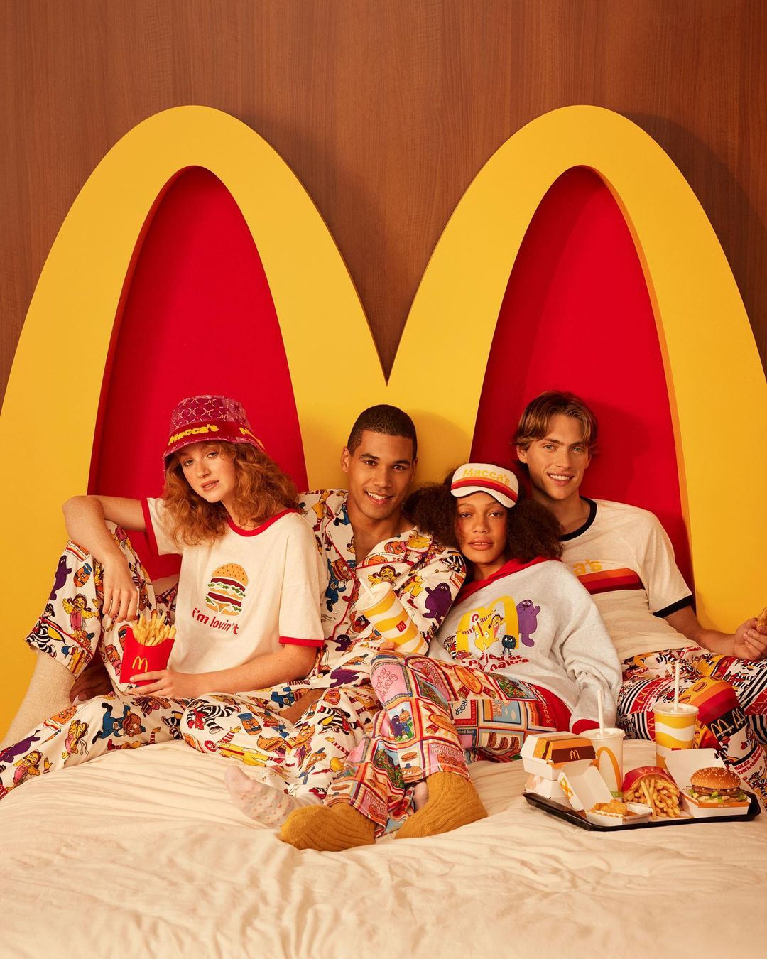 Four young adults in patterned pajamas sit on a bed in front of a golden McDonald's logo, with a tray of McDonald's food.