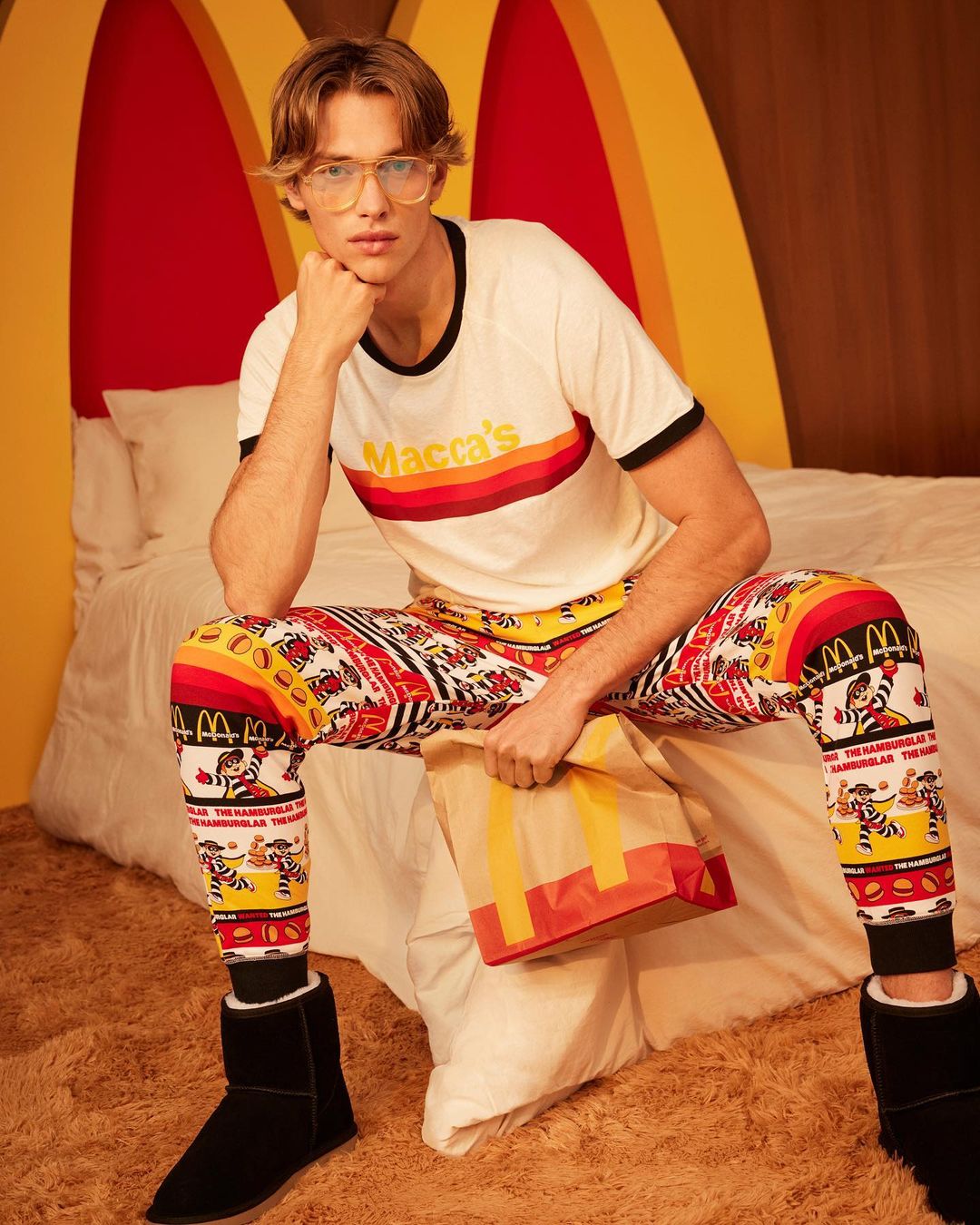 Male model poses on bed with red and yellow backdrop in printed McDonald's themed pajamas, eyeglasses, and slippers holding paper bag.