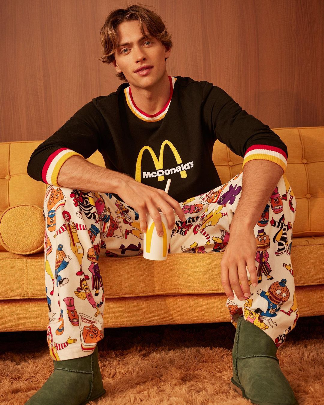 Young man sitting on a yellow couch wearing McDonald's themed pajamas, holding a drink, indoors, with a 70s aesthetic.