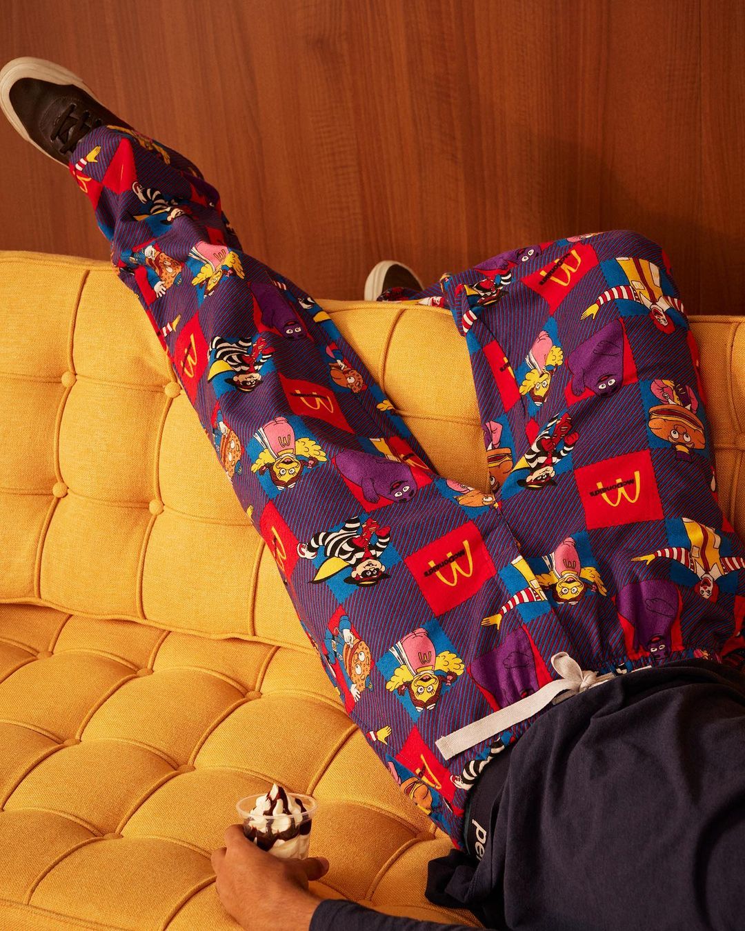 Person lounging on a yellow couch with a dessert, wearing McDonald's themed pants and black shoes, in an indoor setting.