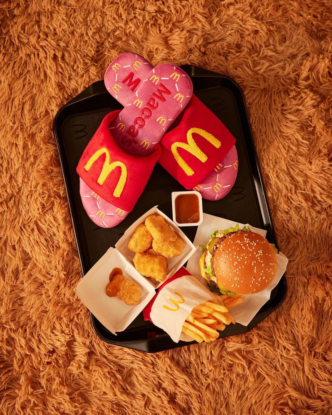McDonald's themed slippers, burger, fries, and chicken nuggets arranged on a black tray on a brown shaggy surface.