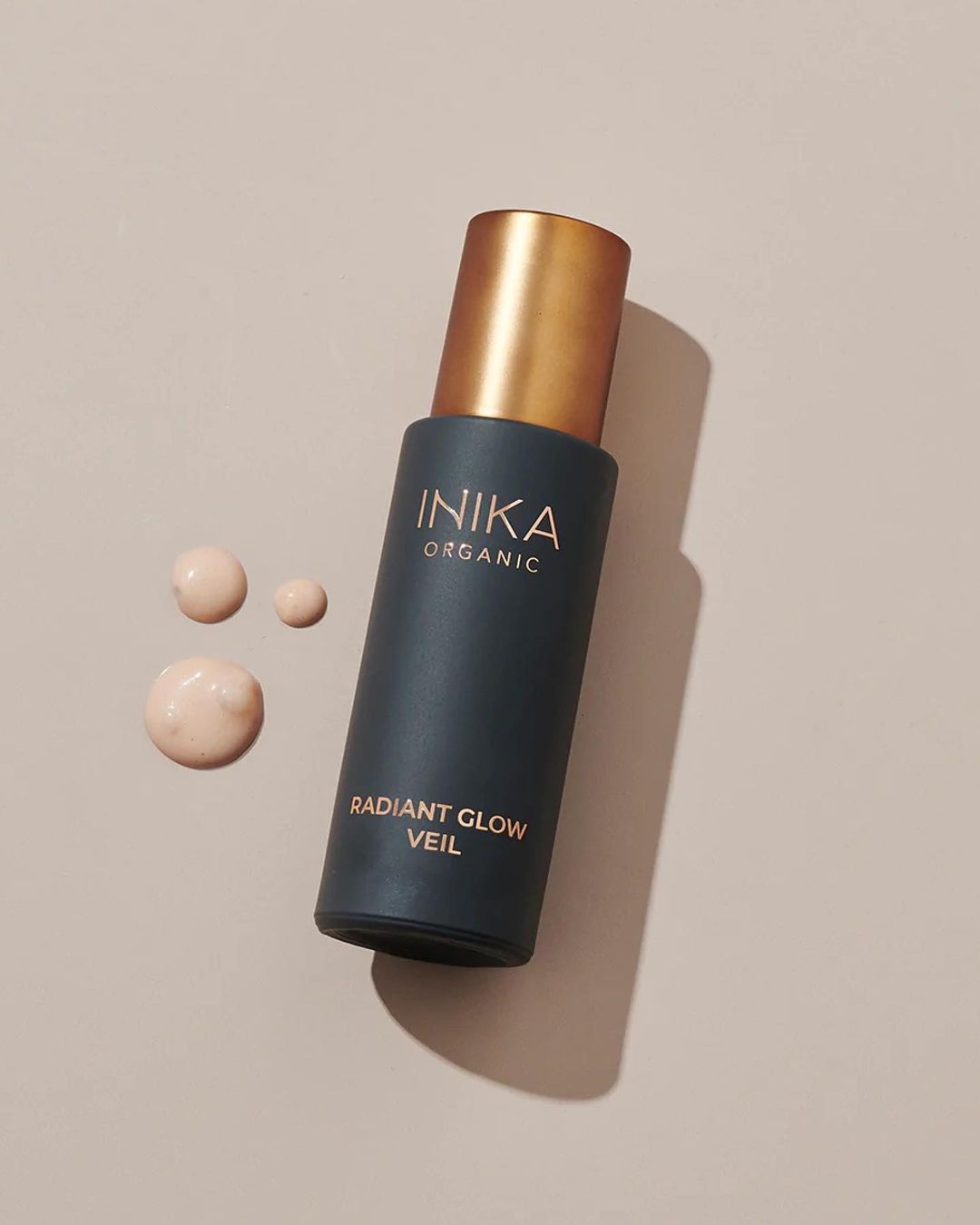 Cosmetic product by Inika Organic, Radiant Glow Veil, displayed indoors with product drops on a beige surface, showcasing beauty item.