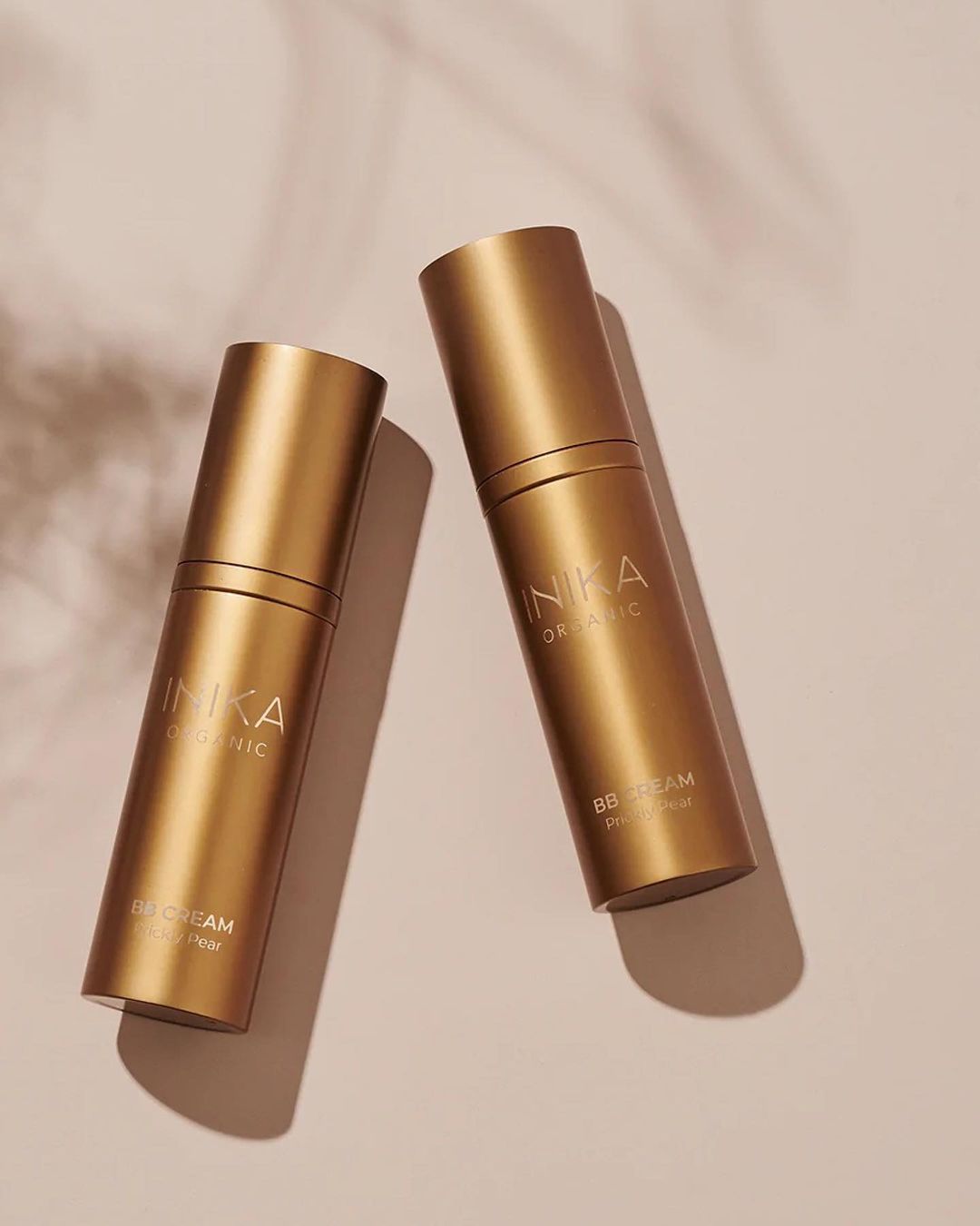 Two gold cosmetic bottles of INIKA Organic BB Cream are angled on a light surface with subtle leaf shadows, conveying beauty.