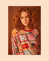 A woman with wavy ginger hair wearing a colorful fast-food printed long-sleeve shirt against a wood-paneled wall.