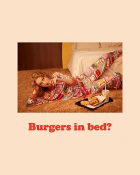 Woman in patterned pajamas lying on a thick brown carpet beside a tray of fast food with a bed in the background.