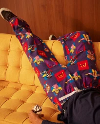Person reclining on a yellow tufted couch wearing patterned purple loungewear pants and holding a small dessert.