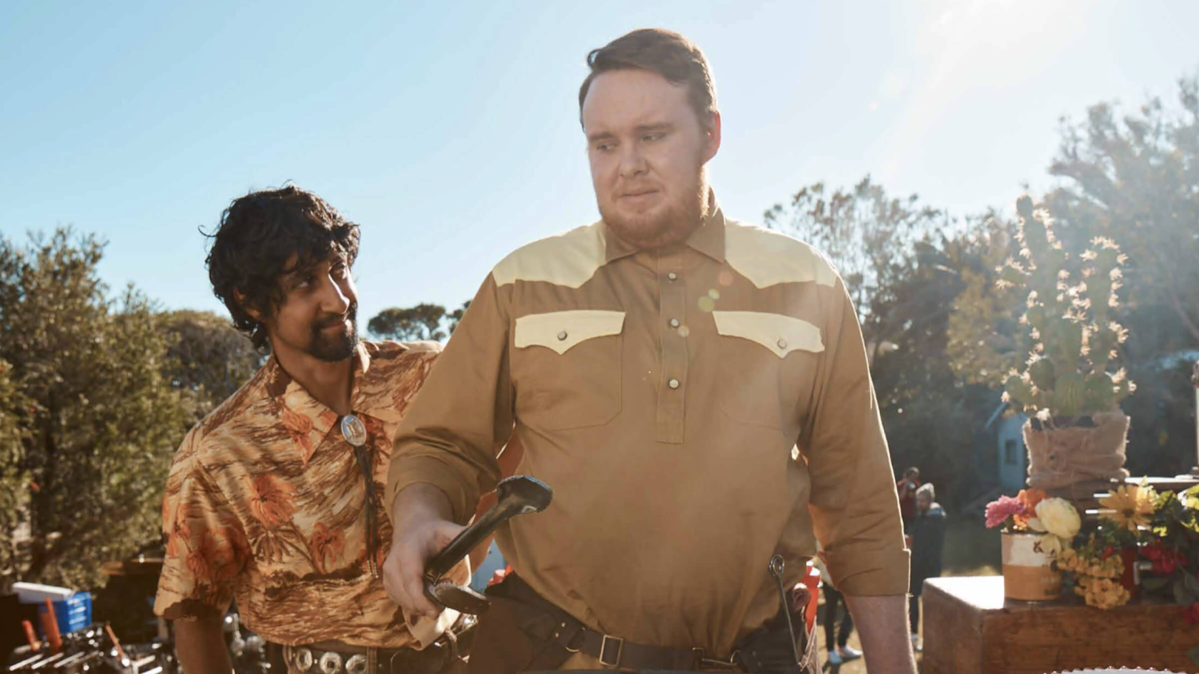 Two men stand outdoors in sunlight; one with a moustache wears a western shirt and looks off frame, the other looks down at the ground.