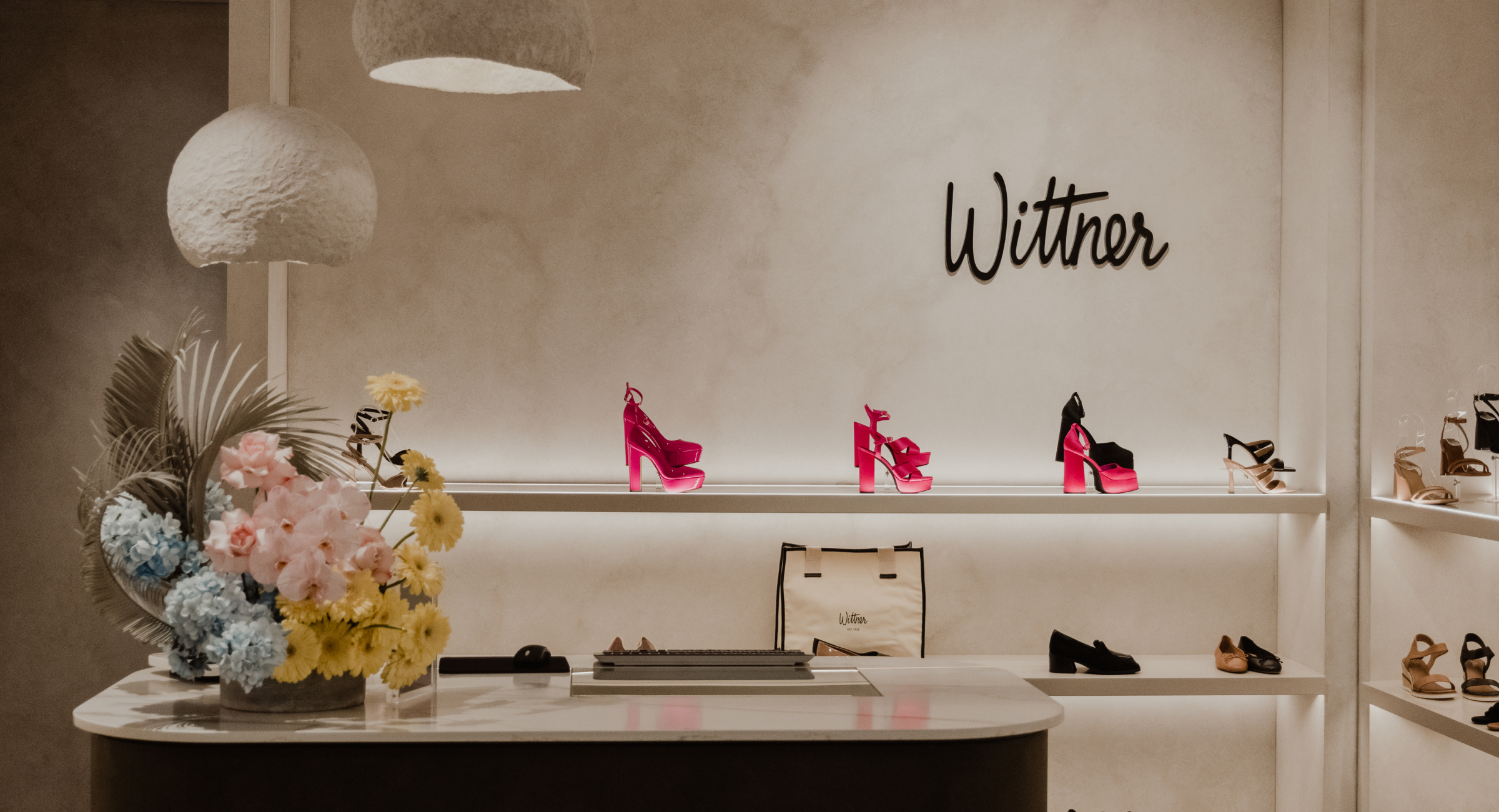 Retail display showcasing pink platform heels on lit shelves against a branded wall with a marble counter and floral arrangement.