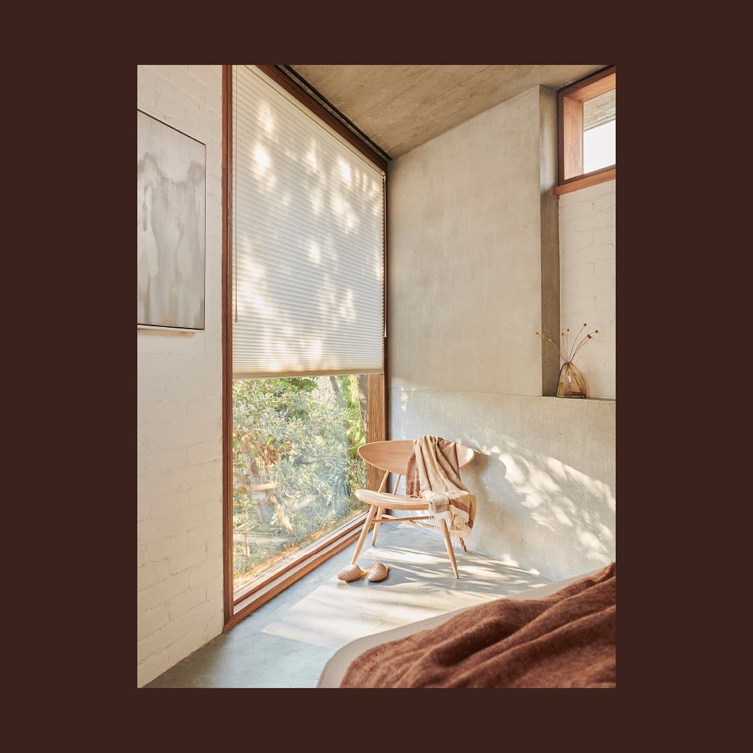 Bedroom interior featuring a wooden chair with a blanket, window with blinds, concrete wall, and view of greenery outside.