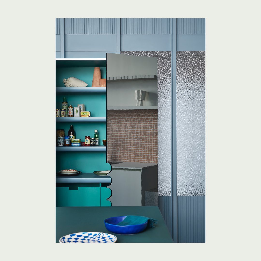 Interior view of a blue-toned room featuring a turquoise shelf stocked with bottles, blue bowls, textured glass, and tiled walls.