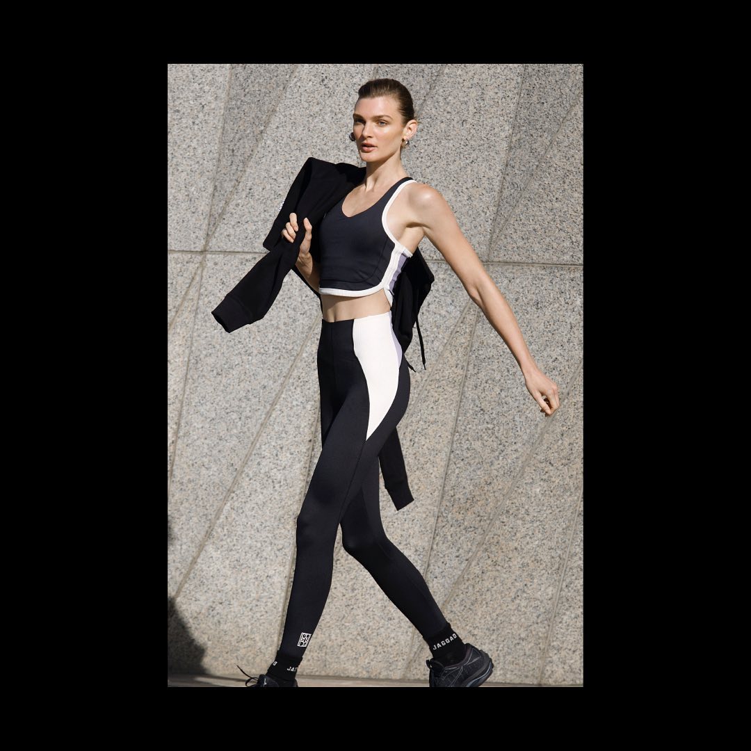 Athletic woman in black-and-white sportswear walking against a textured grey wall, wearing a jacket slung over her shoulder.
